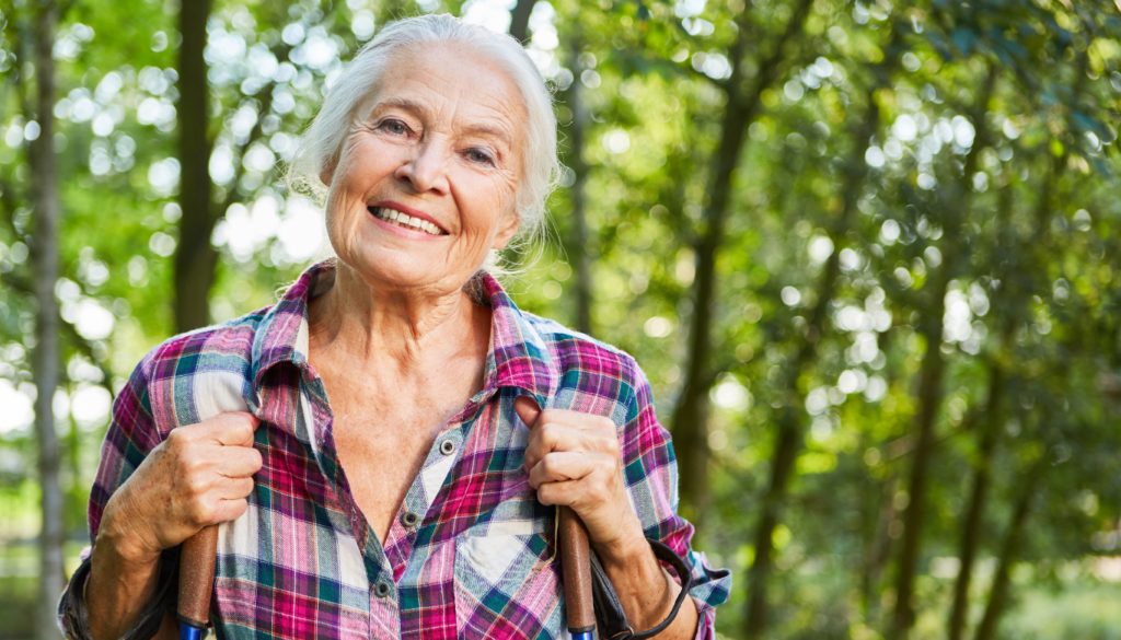 Smiling elderly woman in a plaid shirt outdoors, holding trekking poles in a sunlit forest.