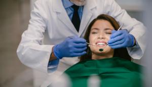 the-dental-project-dental-crowns | The Dental Project | The Dental Project Dentist in a white coat and blue gloves uses dental tools to examine a patient's teeth while the patient rests in a chair with a green bib.